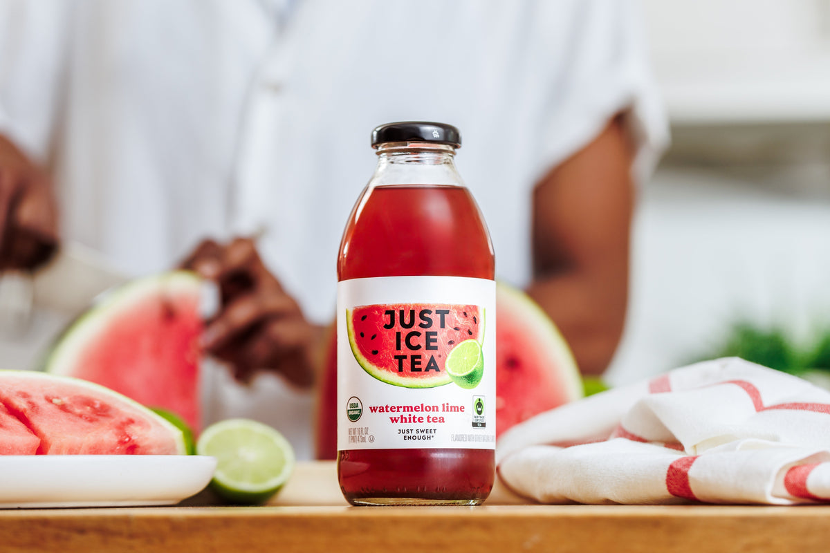 Front view of Just Ice Tea watermelon lime white tea bottle with watermelon and lime fruit on a kitchen table